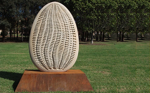 Oneness sculpture - displayed outdoors on a grassy area with trees in the background. The scupture is egg-shaped and off-white in colour with patterns running from top to bottom. It sits on a brown stand.