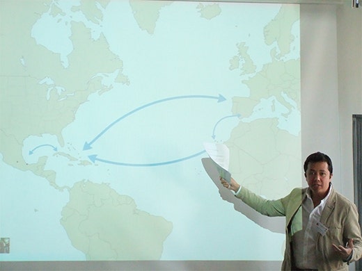 Alejandro Miranda Nieto giving a presentation - gesturing at a slide on a screen which shows a map of the world and arrows between Mexico, Europe, and across the North Atlantic Ocean.