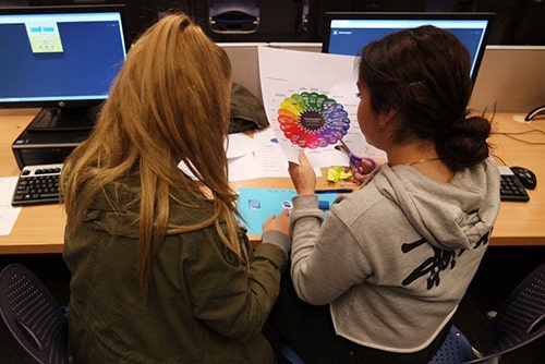 Two girls sitting at a table next to computers making a collage.