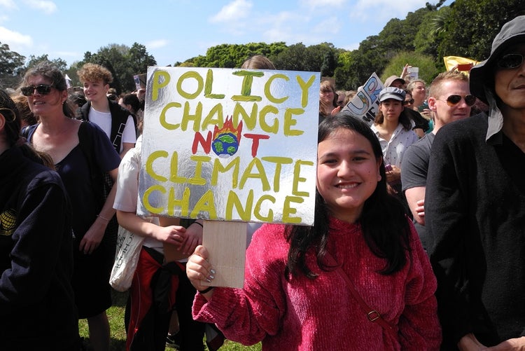 A young person in amongst a crowd holds a sign saying 'policy change not climate change'.