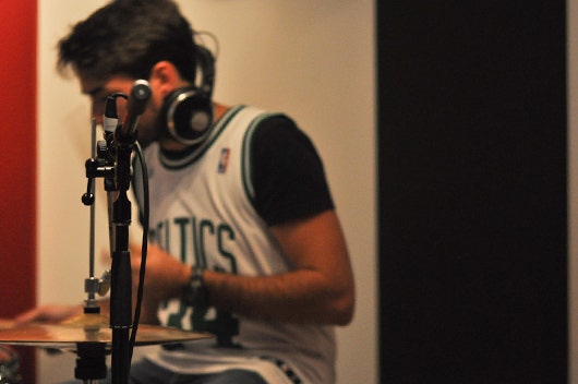 A young man in a recording studio - a microphone and drums in the foreground. He sits in the background with headphones on, out of focus.