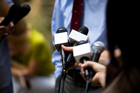 The hands of journalists holding microphones towards a man in a shirt and tie (face not visible).