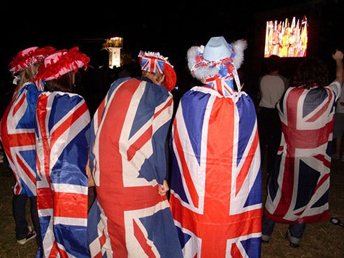 The backs of people who are wrapped in the UK flag, as they stand watching the closing ceremony on a TV screen in a park.