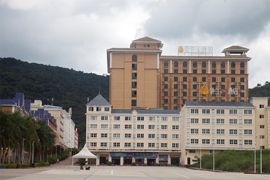 Buildings and an empty parking lot stand in front of tree-covered hills.