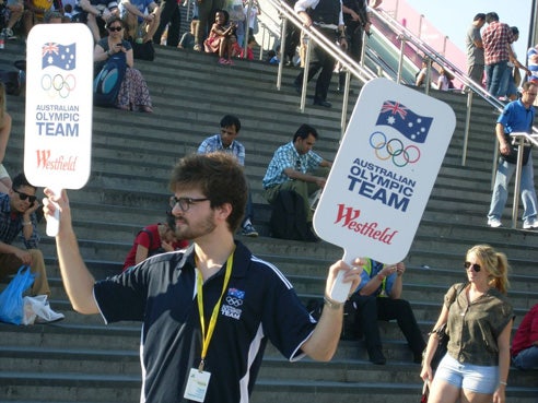 A man stands on the stadium steps holding a sign in each hand - 'Australian Olympic Team Westfield'.