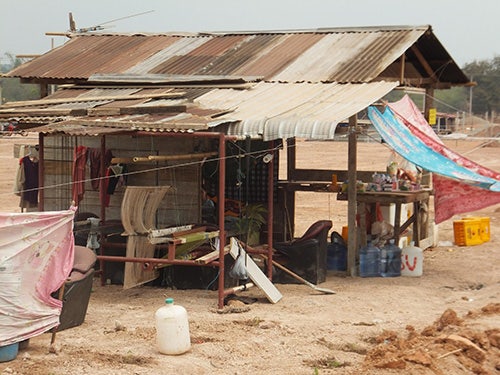 A shelter constructed out of wood, with a tin roof. Part of it is enclosed. Sheets hang from the roof. Underneath the roof are clothes, a table, water bottles, and other possessions.