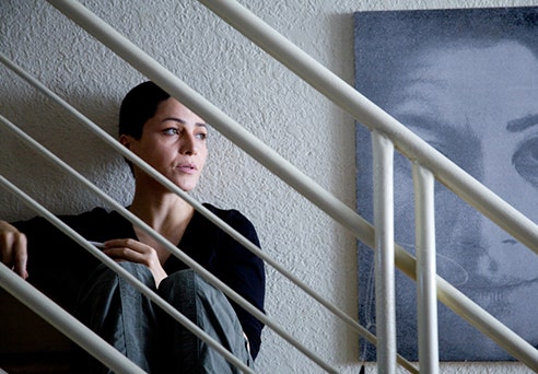 A woman sits on a staircase. On the wall behind her is a painting of another woman's face.