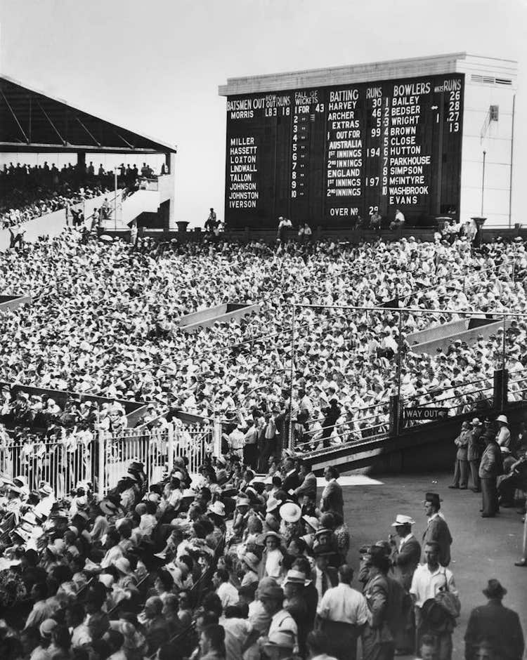 A black and white scene of dense crowds gathering in front of a cricket scoreboard in 1950