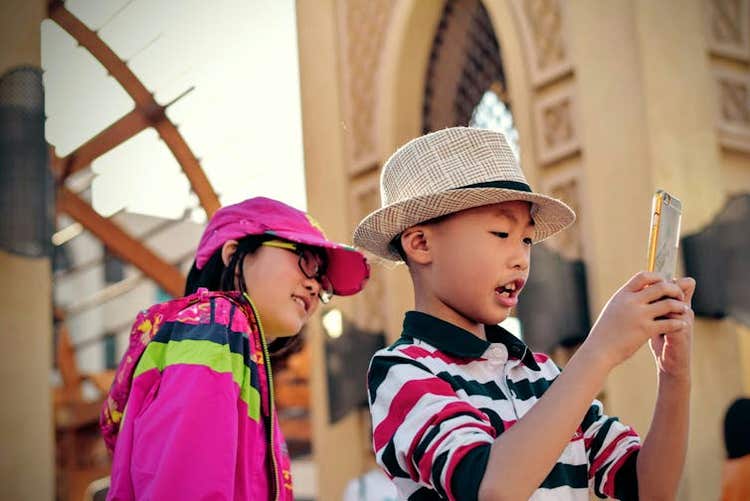 Two kids outdoors in bright clothing taking a photo with a smartphone.