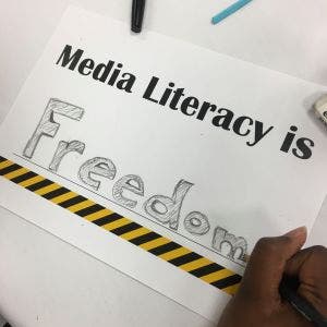 The blank space on a worksheet reading ‘Media Literacy is….’ has been filled out with the word ‘Freedom’ in strong, bold lettering. A hand holding a pencil is shown in the lower right corner putting the finishing touches on the word.