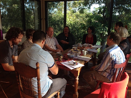 A group sits around a wooden table in a sunroom with trees showing through the full-length glass windows.