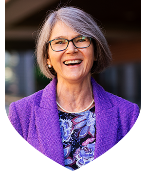Portrait of a person with short grey hair wearing glasses, a purple blazer and a patterned top.