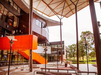 Contemporary campus courtyard featuring glass-fronted buildings, covered outdoor spaces, seating areas and a prominent orange staircase.