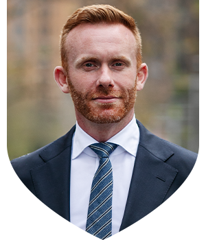 Man with red hair and beard wearing a navy blue suit jacket and tie with a white shirt facing the camera.