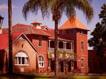 Historic brick building featuring a red tiled roof, tower, verandah and palm trees surrounded by gardens.