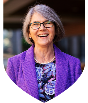Portrait of a person with short grey hair wearing glasses, a purple blazer and a patterned top.
