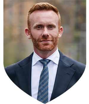 Man with red hair and beard wearing a navy blue suit jacket and tie with a white shirt facing the camera