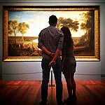 A man and a woman look at a landscape painting, with their backs to the camera.