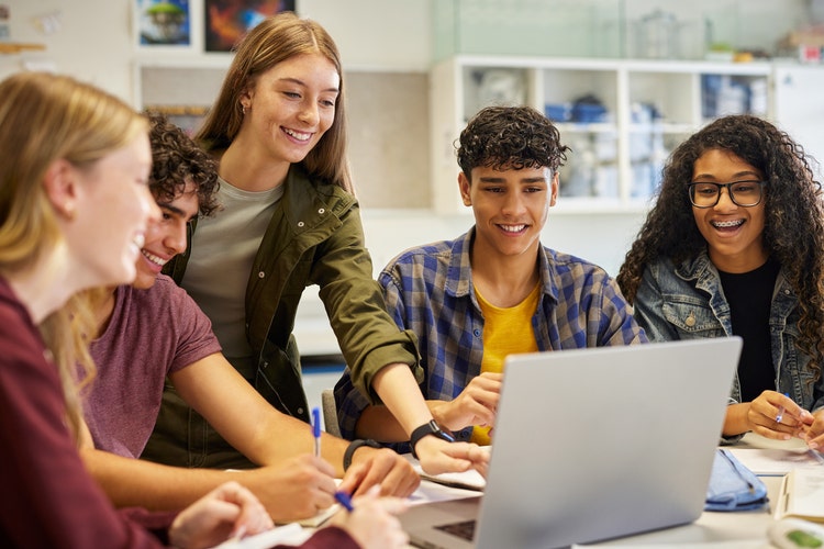 Multiethnic casual students using laptop during research, working together on a project. Diverse teenagers pointing at screen while engaging in collaborative learning. Group of happy classmates sharing ideas and taking notes during a digital study session for exam.