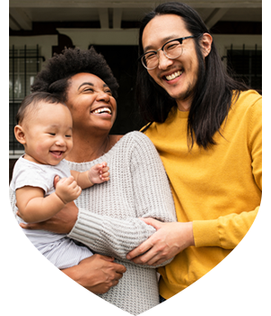 Two adults standing together and holding a smiling baby. One adult wears a light grey knitted sweater and the other a mustard-yellow jumper.
