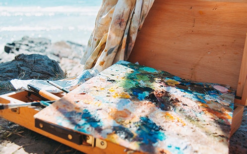 An abstract painting on canvas and a wooden box of art supplies sits on the rocks near the ocean.