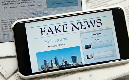 A mobile phone with a fake news headline displayed on the screen, sitting over a tablet and newspaper.