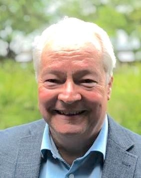 photo of Kurt, smiling at the camera, fair skin, white hair, blue shirt and jacket, grass and trees in background