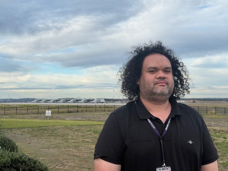 photo of person with jaw length black curly hair, short whiskers, wearing a black polo shirt, they are standing outside with grass in the background