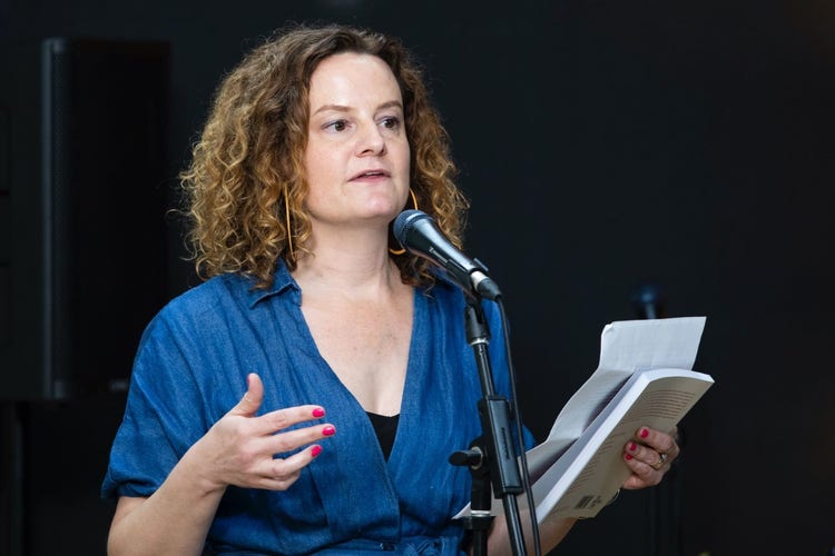 Felicity Castagna wears a blue shirt and holds a sheet of paper and book in her left hand. She is standing in front of a microphone, and speaking to an audience off screen.