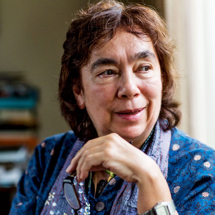 Alexis wears a blue and cream spotted shirt, with a light purple scarf. Her left hand is posed right below her face and she looks off into the distance.